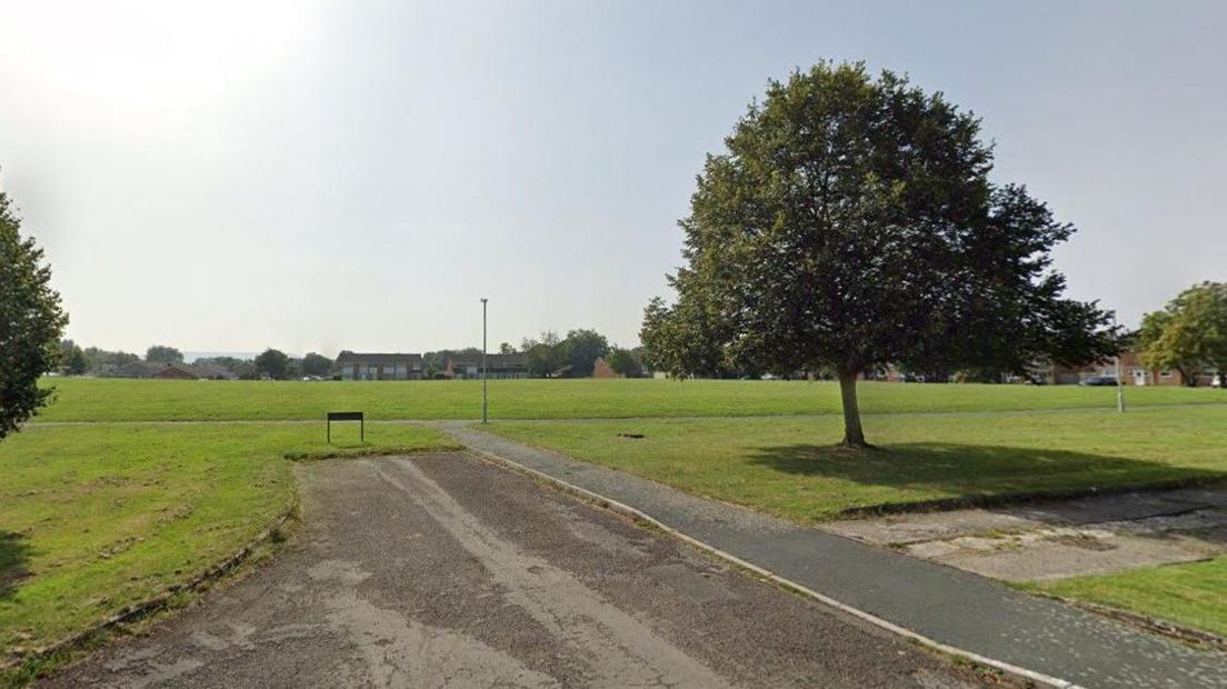 A stretch of green playing field in Foxbridge with a few trees and houses in the distance.