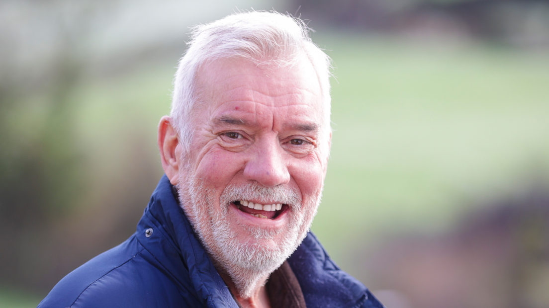 A man smiles into the camera. He's wearing a navy coat and a brown top. He's outside. The background is blurred but he's outside.