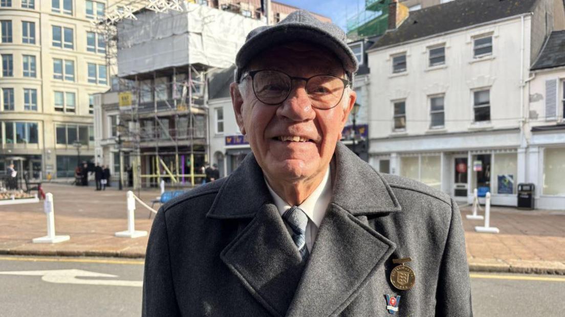Brian Biddle is seen smiling at the camera in a grey coat, white shirt and tie. He is wearing a cap and glasses. His medal is gold.