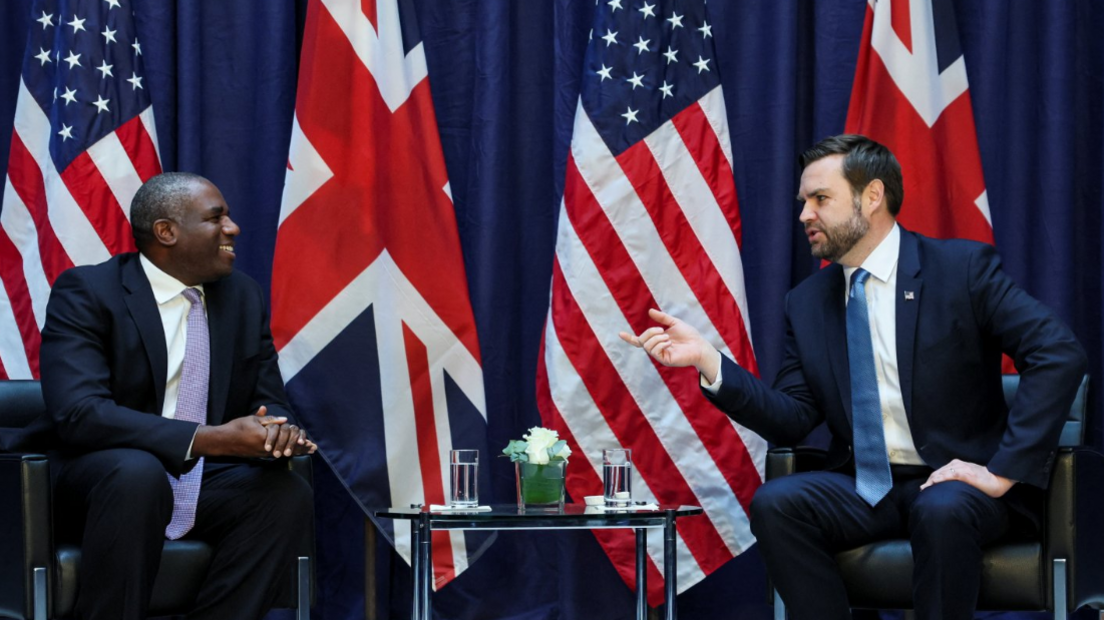 David Lammy smiles across a table at JD Vance, who is talking and gesticulating at a meeting. The backdrop is large UK and US flags. 