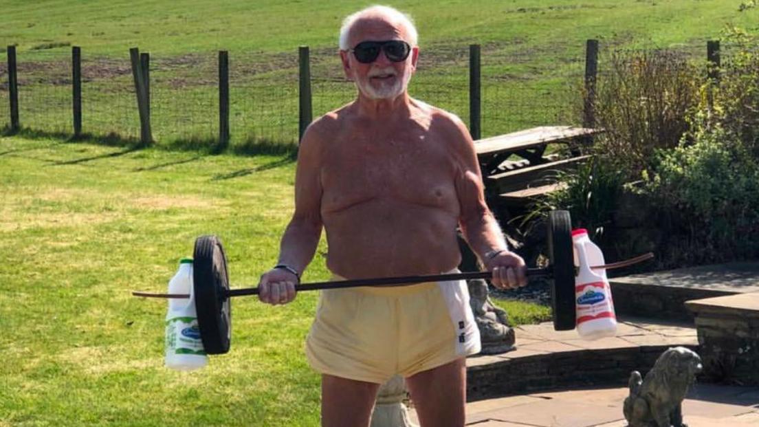 Mick Wadge stands on a patio holding a homemade barbell weighted with wheels and milk containers