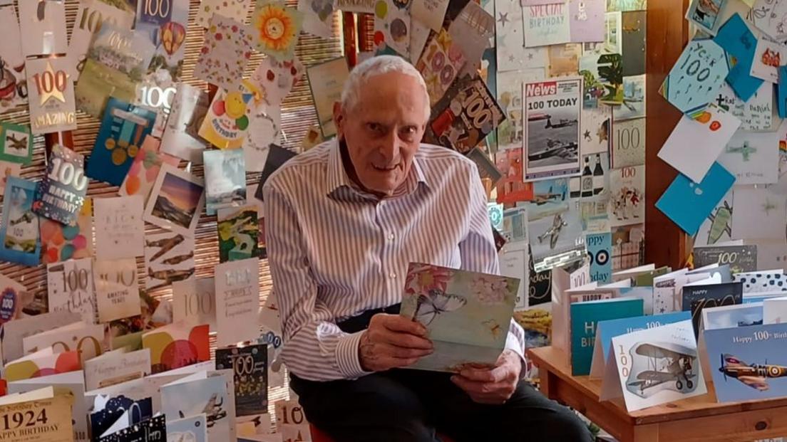 An old man in a white shirt and black trousers holds a card and smiles at the camera, in a room that is filled with birthday cards - on the floor, on tables, and covering every inch of the walls