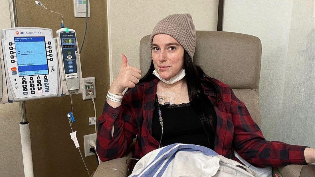 Molly gives a thumbs up to the camera as she's hooked up to a medical machine. She is sat on a chair waring a beanie and plaid shirt with her long dark brown hair loose around her shoulders.