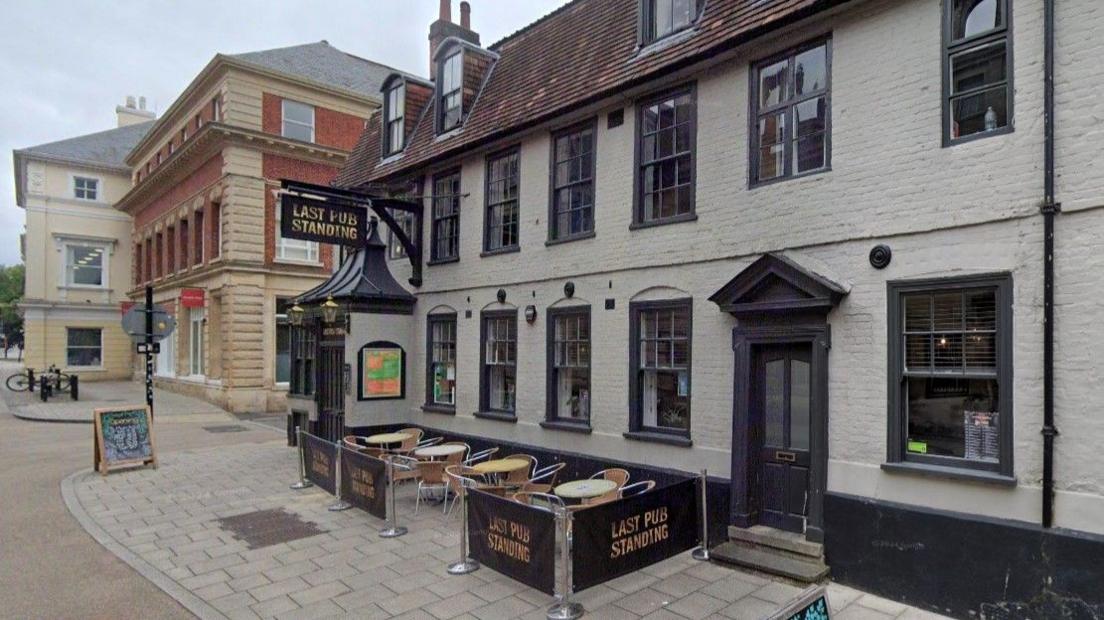 A pub with grey walls and dark grey window and door frames is shown with its name sign above the door to the left and an empty seating area in the centre of the frame.