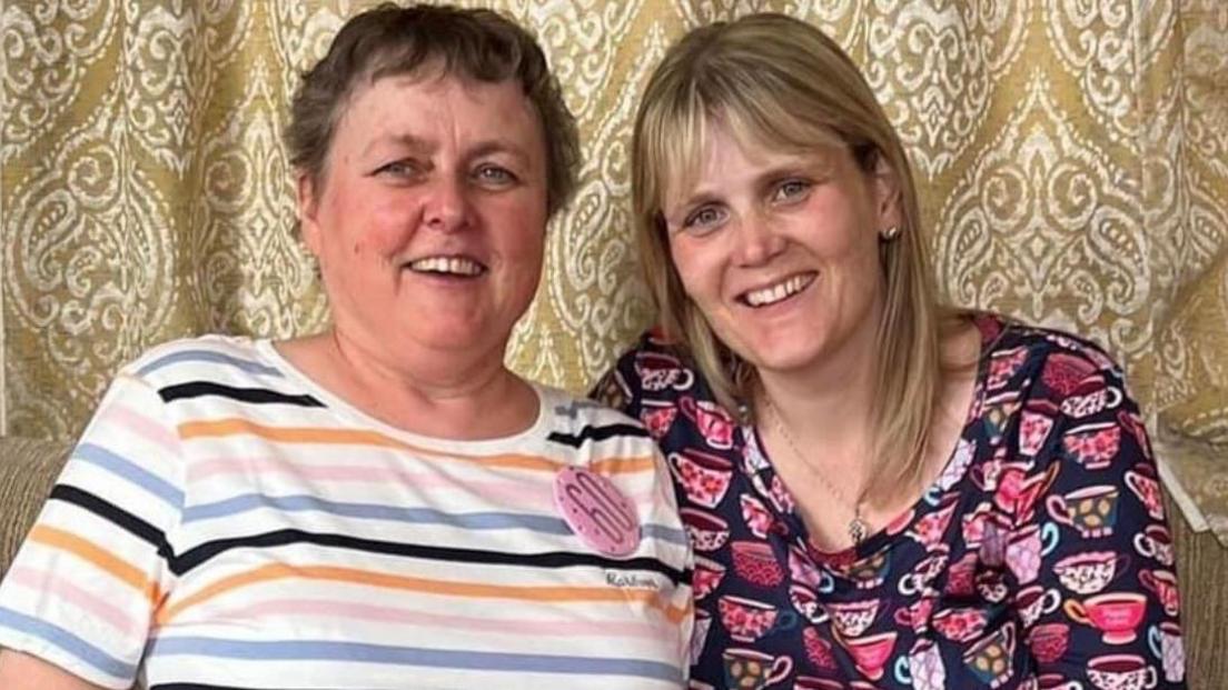 Cheryl Woods wearing a stripy top with a pink 60th birthday badge. She has short mousey brown hair. On the right is Sarha Smith, who has medium-length blonde hair and is wearing a long sleeved top with pink teacups.