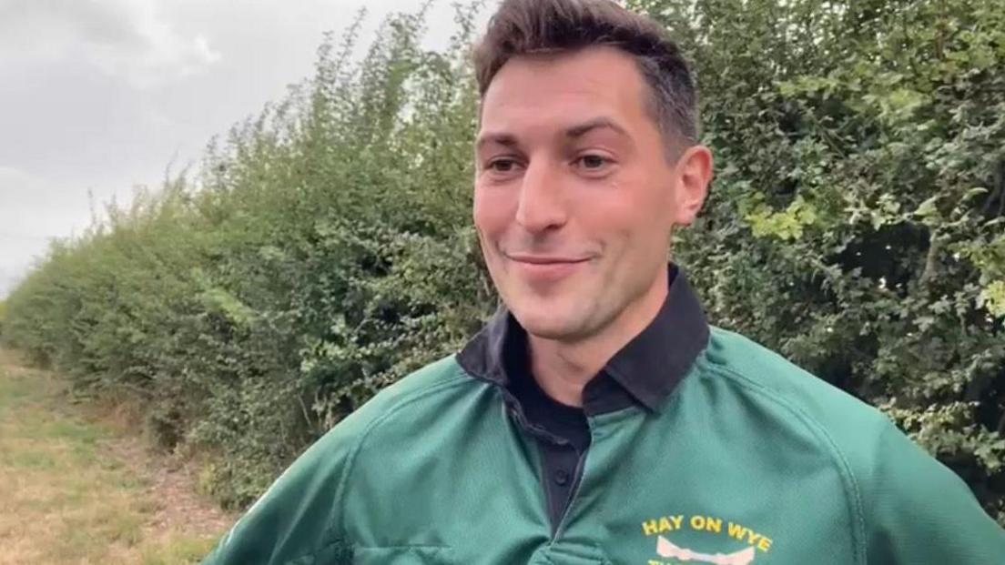 A man with dark hair is wearing a green top. Bushes are behind him and there is some grass on the left of the photo.