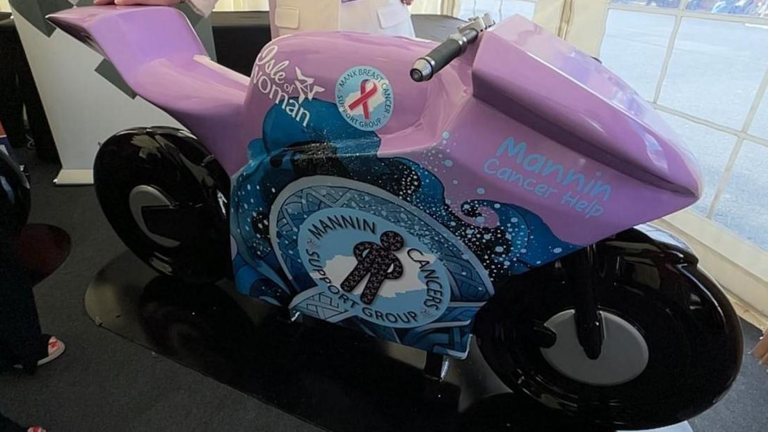 A purple and blue bike sculpture with Mannin Cancers Support Group's logo is on the right.