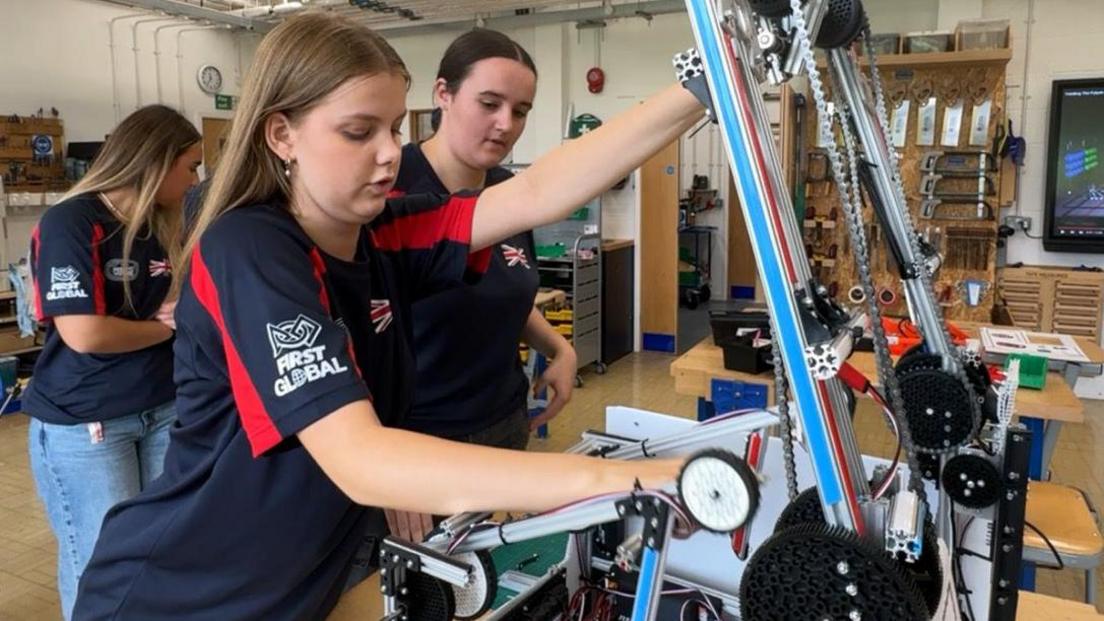 Two girls working on a robot - the device is made up of cogs, chains and mechanical arms