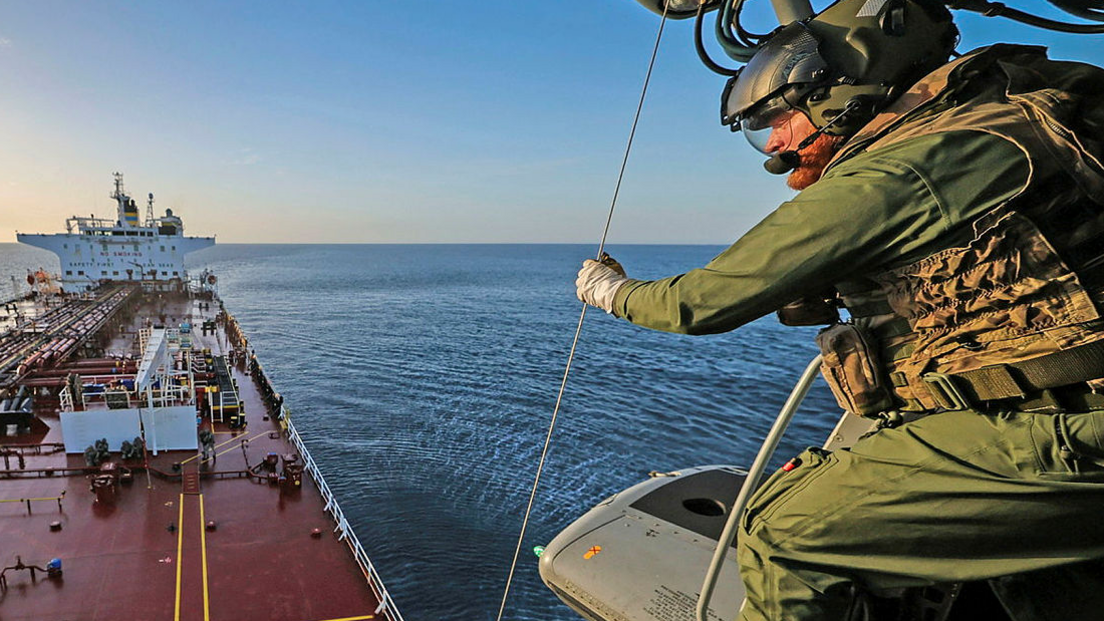 A still showing a military personnel on a helicopter above a large vessel. He is wearing green tactical gear and a helmet and gloves and is looking down at the boat. The water and sky are blue. 