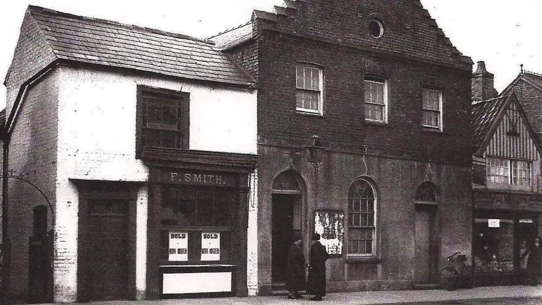 A black and white photo from around 1933 shows the frontage of shops and the church hall, run by St John's Church in March. The building in the middle and a white one to the left were demolished to erect the current former NatWest building, now a Turkish restaurant.