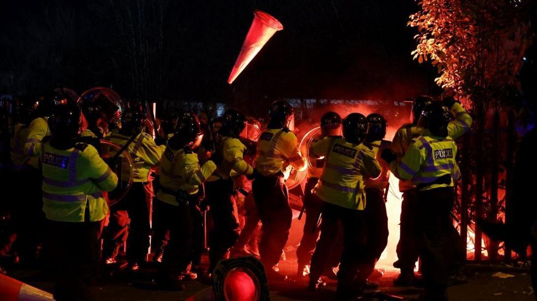 Legia Warsaw fans let off flares as they clash with police officers outside the stadium before the match. Police officers are lined up in hi-vis jackets and there is a glow from the flares. A fence and a tree are visible to one side, lit up in orange.