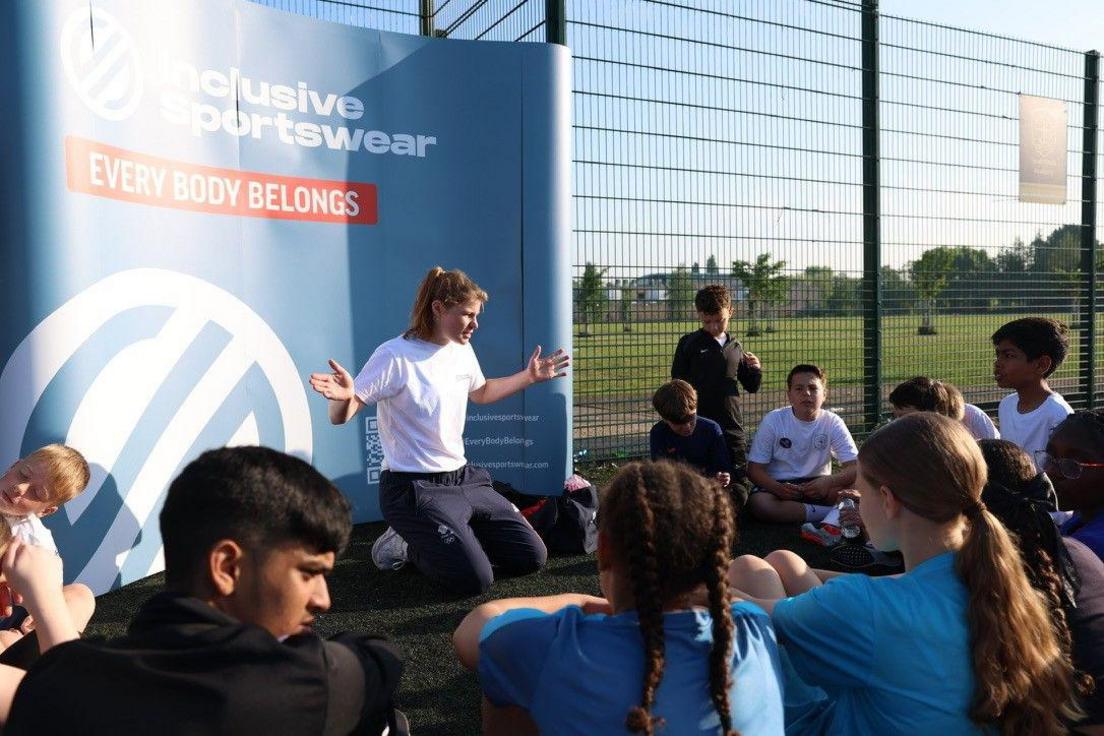 Tess Howard, wearing a white T-shirt, long navy sports trousers and white trainers, sits facing a group of school-aged children gathered around her. She is speaking to them in front of a blue banner with the words 'Inclusive Sportswear : Every body belongs'.