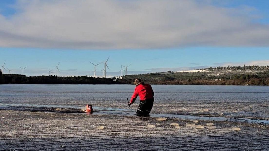 Frankie, with a swimming cap and goggles, swims through a channel in the icy Sweethope Lough. Coach Fenwick Ridely is standing in the water, knee deep, in a red coat and fluffy hat and monitoring her progress. 