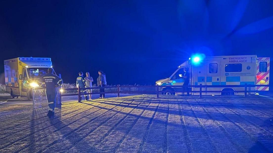 Two ambulances at night with blue light on. A paramedic is standing with two people by a fence and another paramedic is standing by a gate.