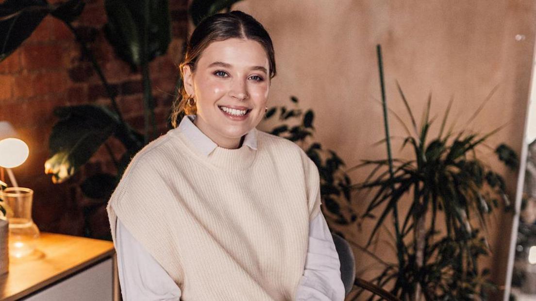 Isobel wearing a white shirt and cream jumper smiles directly at the camera