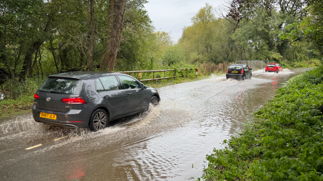 Three cars driving through floodwater on a country road. The water looks to be several inches deep. Either side of the road is greenery, bushes and trees.