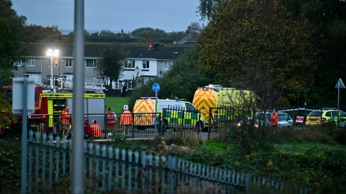 A wide shot of emergency vehicles from the vantage point of a road behind the bridge.  Houses can be seen in the background.  Police tape can be seen to the right, presumably near the river.