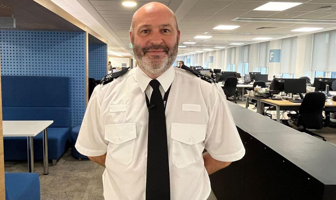 Sgt Stephen Evans is in his police uniform, of a white shirt and black tie. He has a short grey beard and and has his hands behind his back, while standing in a large office with computers in it