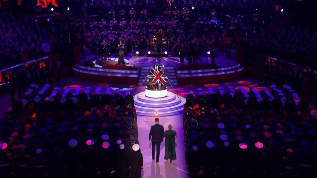 A man and a woman walk down the aisle of the Royal Albert Hall towards a Union Jack flag.  Flanked by audience members in military wear.