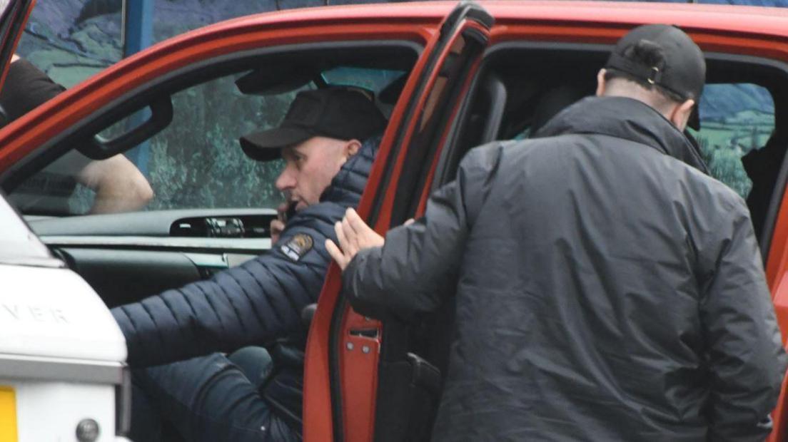 Image of Arsen Feci in the front passenger seat of a red car. There is a another man with his back to the camera with the back-left passenger door open.