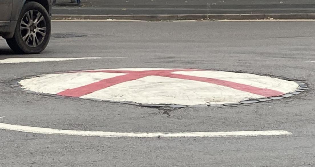 A red cross can be seen on a white mini roundabout, with arrows around the middle. The wheel of a car can be seen as it is driven round the roundabout.