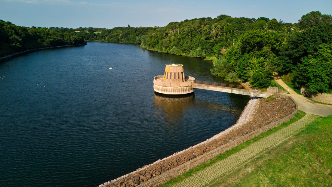 There is a large expanse of water in a reservoir with green shrubs and trees beside it and a field and path in front. There is a bridge over the reservoir to a concrete valve tower.