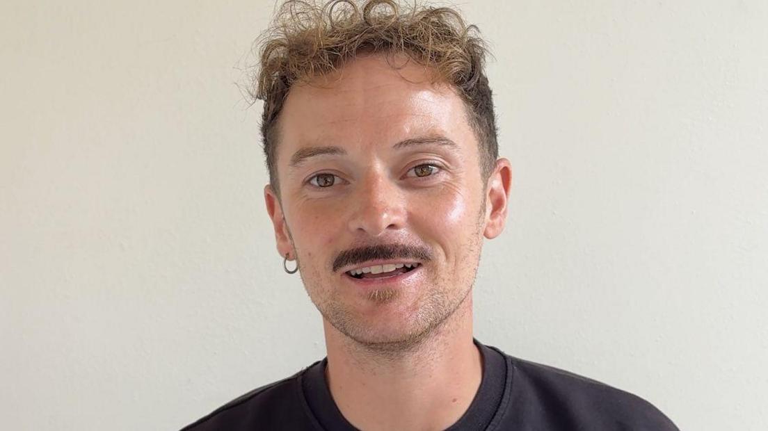 Tom is wearing a black tshirt and is standing in front of a beige wall.