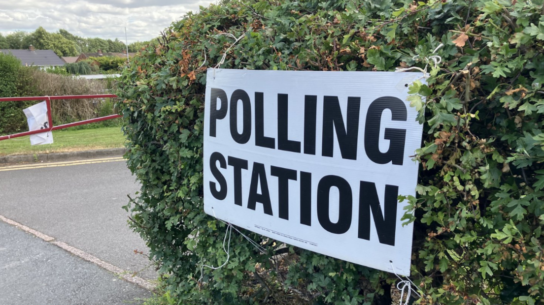 A white sign attached to a hedge reads POLLING STATION in blocked capital black lettering.