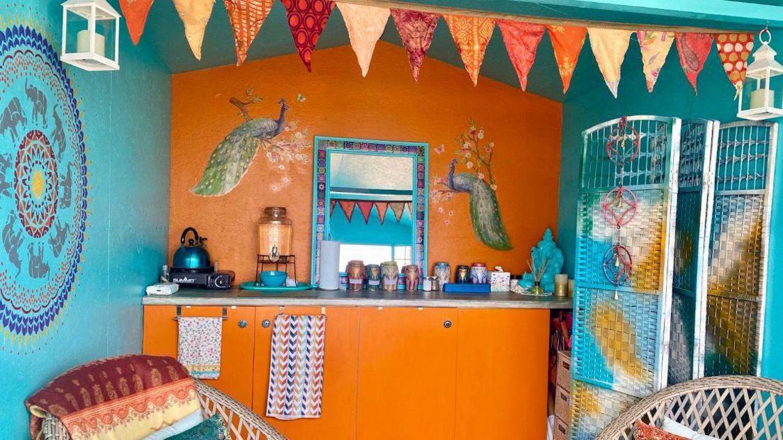 The interior of the beach hut has turquoise walls on both sides and the back wall is orange with orange cupboards. Bunting is hanging in the middle and there are two wooden chairs. Artwork has been painted on the walls, including of two peacocks, and there are various tea towels, a kettle, a mirror and pots neatly lined up.