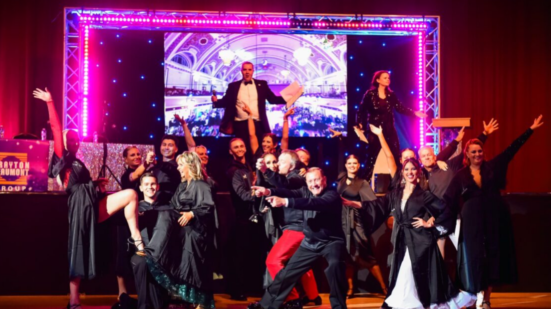 More than a dozen people in smart clothing are stood in front of a stage, with a number of them throwing their arms in the air. There is a brightly lit frame on the stage, with a man and woman looking out towards the audience