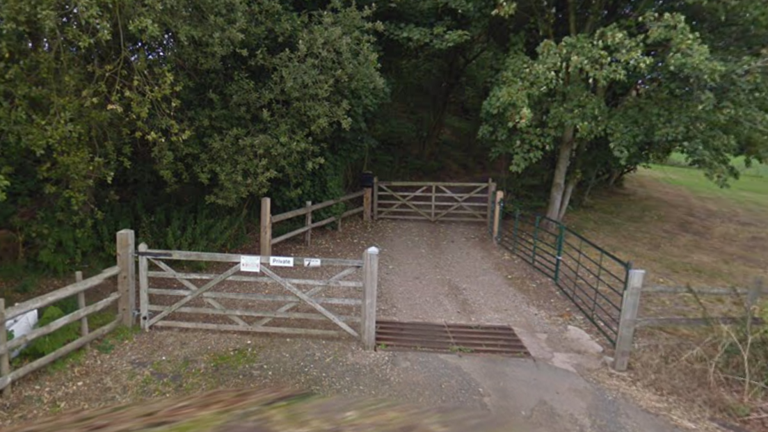 The entrance to a property with wooden gates and fencing, with trees to the left and a field to the right