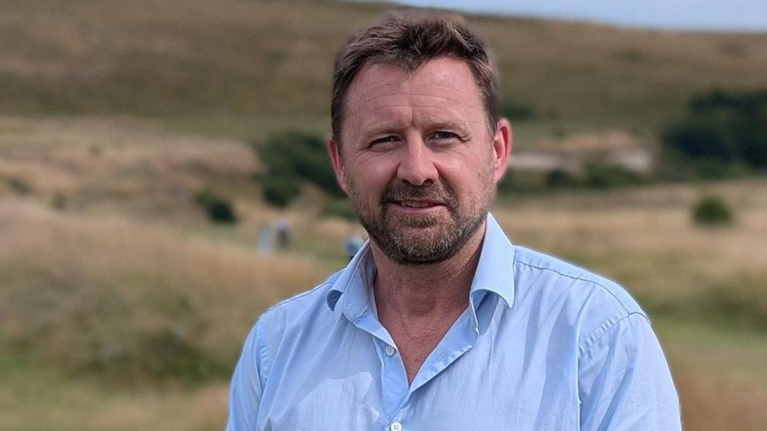 Siôn McGeever, chief executive for South Downs National Park, wearing an open button blue shirt and stood in a green field area.