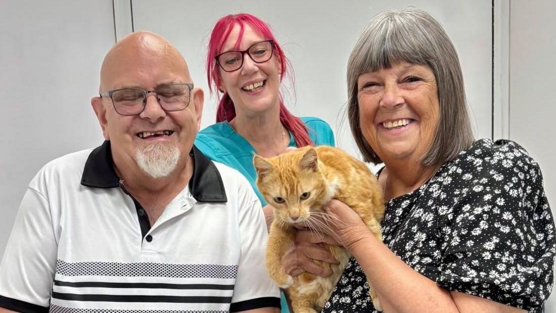 Robert Poile, in a black and white top, and Debbie Poile, wearing a black and white dress, are at the vets holding their ginger cat Primrose, with veterinary nurse Chris Guscott behind them.