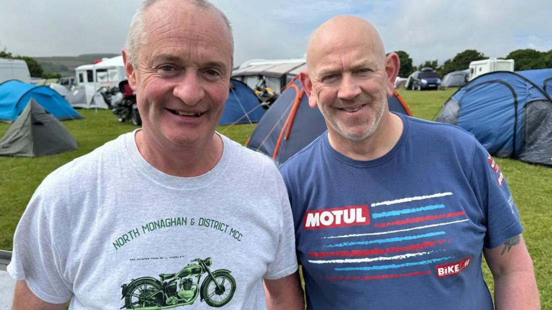 Paddy Corey and John Little stand in field of tents. Paddy wears a grey t-shirt with a green motorcycle on it, while John wears a blue T-shirt with white, blue and red stripes.