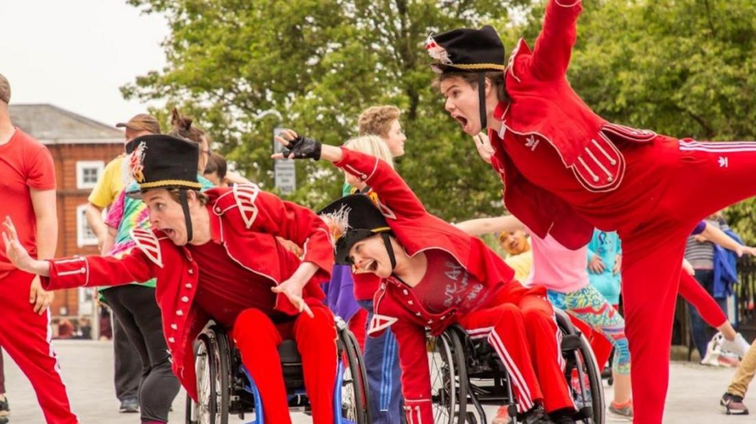 A man and a woman in wheelchairs and a standing man, all from Stopgap Dance, lean towards the Forum in red costumes as part of a show called Frippery for the Norfolk and Norwich Festival in 2019