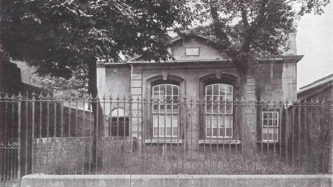 An archived image of Pile Street School in black and white. It is a modest building with two large windows at the front, with a smaller one on the right and an arched doorway on the left. There is a walkway leading up to the door and a grassy patch and trees outside the front of the building, separated from the road by spiked railings.