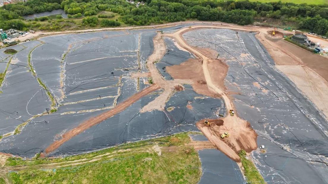 An aerial view of a former landfill site which has been mostly covered in a dark material. Several vehicles can be seen parked on the site. A number of trees are visible at the top of the frame.
