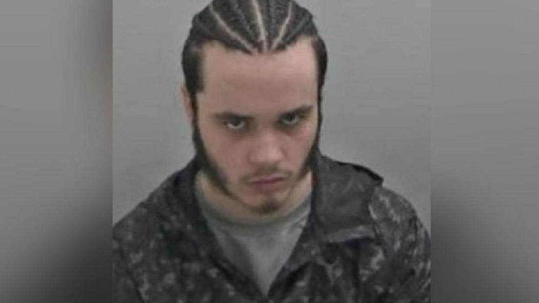 Zach Pearson in a police mugshot. He has braids in his hair and he has his head bowed but is looking at the camera.