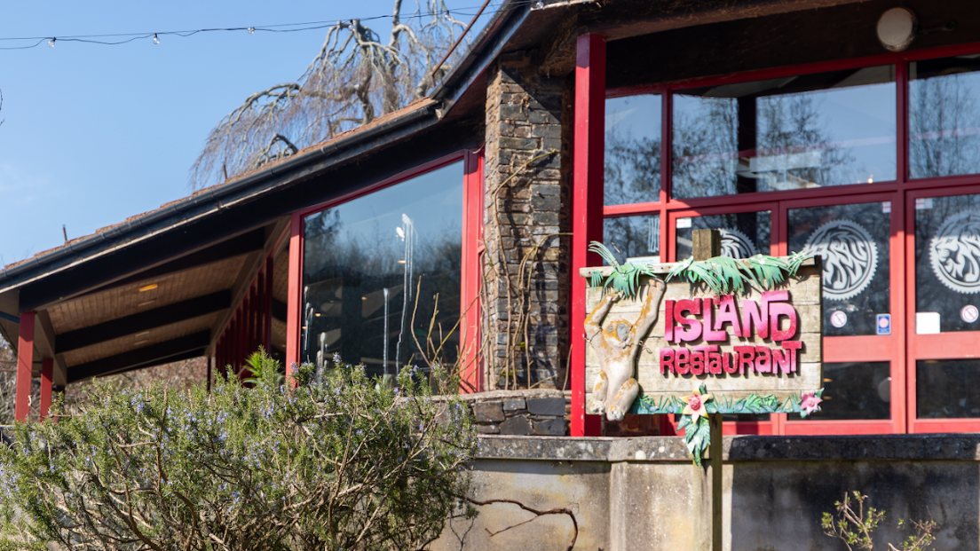The picture shows the exterior of a building labelled "Island Restaurant." The structure features large windows with red frames and a stone facade. In front of the building, there are bushes and trees, adding greenery to the scene. The restaurant sign is decorated with tropical-themed elements like leaves.