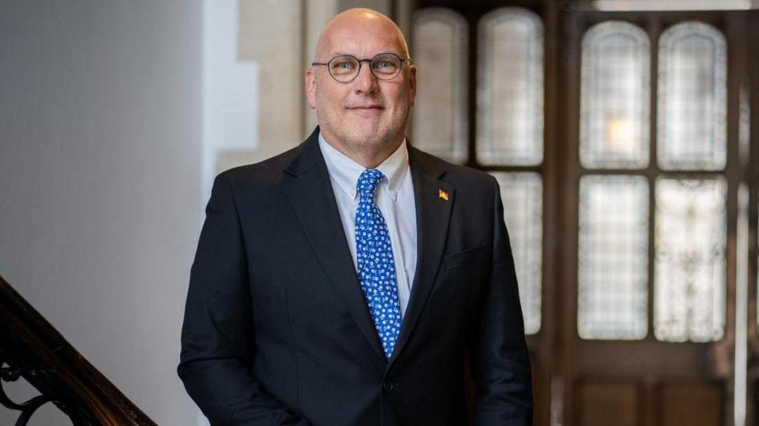 Councillor Nick Adams-King stands at the bottom of an ornate wooden staircase in the Hampshire County Council offices. The background is blurred, but it is a set of wooden doors with four sections of stained windows. Cllr Adams-King looks directly at the camera, he is wearing round black framed thin wire glasses, with a deep blue blazer, a white shirt underneath and a paler blue tie with a white floral details. He has a golden flag pin on the right panel of his blazer collar.