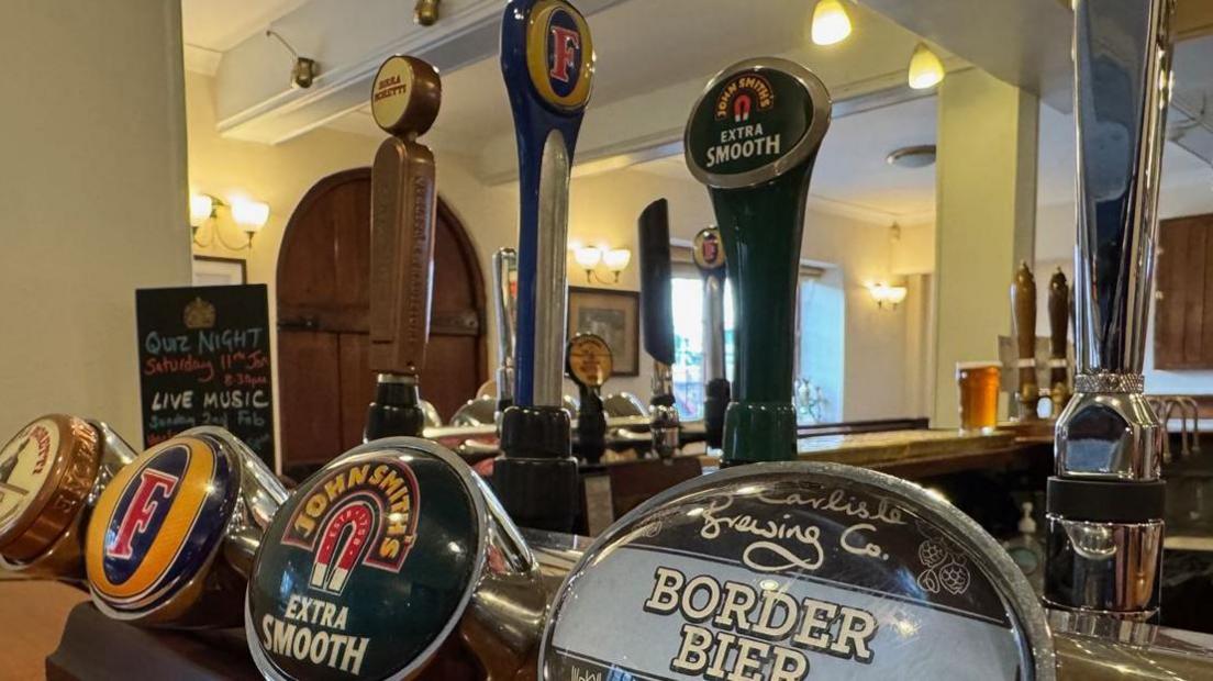 View of four beer pumps on a bar. Labelled from right (the closest) to left; Carlisle, John Smith, Fosters, Moretti. Behind them, at a right angle are more beer pumps, a pint on the bar, and a window