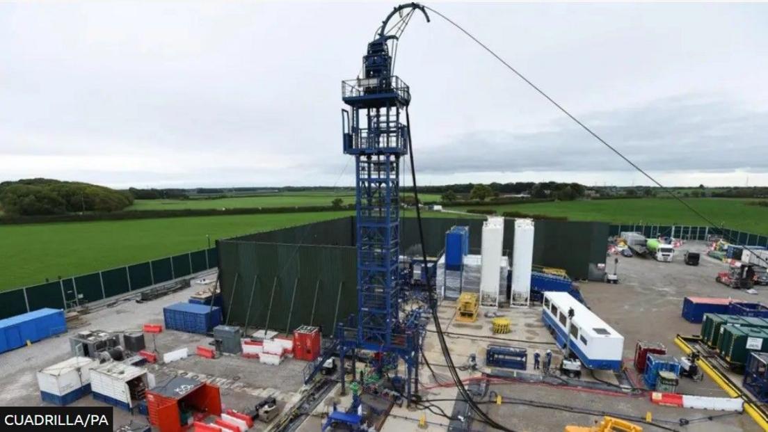 Fracking equipment and various storage units can be seen on the site, which has green fencing all around.