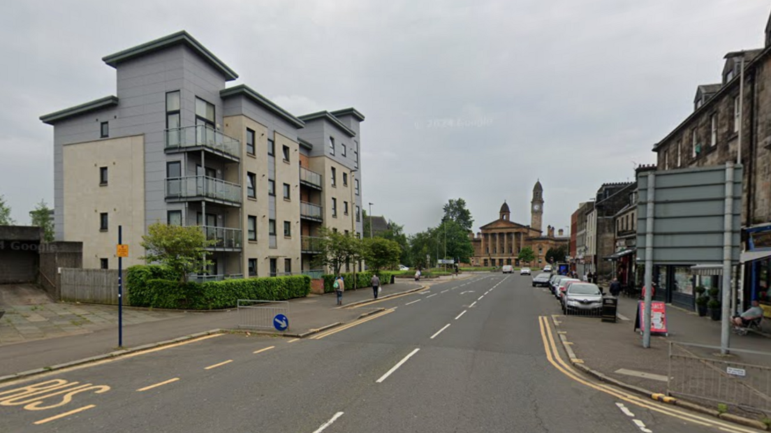 A road filled with parked cars on one side, a bus lane on the other and various homes and shops on either side 