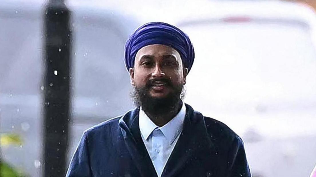A man with a dark beard wearing a blue Sikh turban, a white shirt and dark jacket.