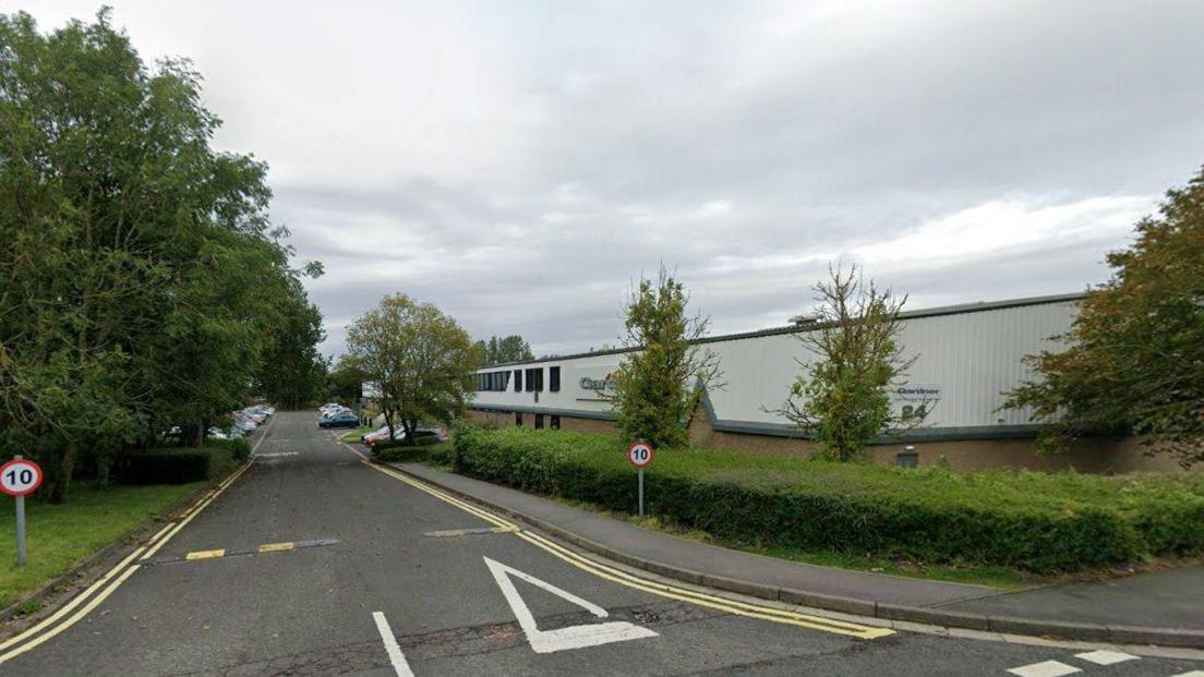 A factory building sits on the right hand side of the road lined with bushes and trees.