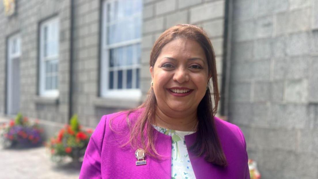 Munazza Malik wearing a bright purple jacket with a floral white t-shirt beneath her jacket. She has long brown hair and has been photographed in front of a grey stone wall. 