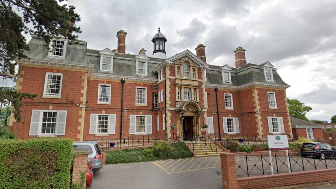 Astell House Care Home in Cheltenham. It is a red brick building with lots of windows. Cars can be seen parked in front of it. 