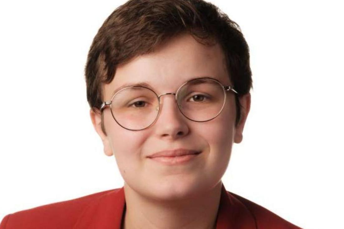 A professional headshot of a young woman with short cropped dark hair and round glasses. She is wearing a red blazer. 
