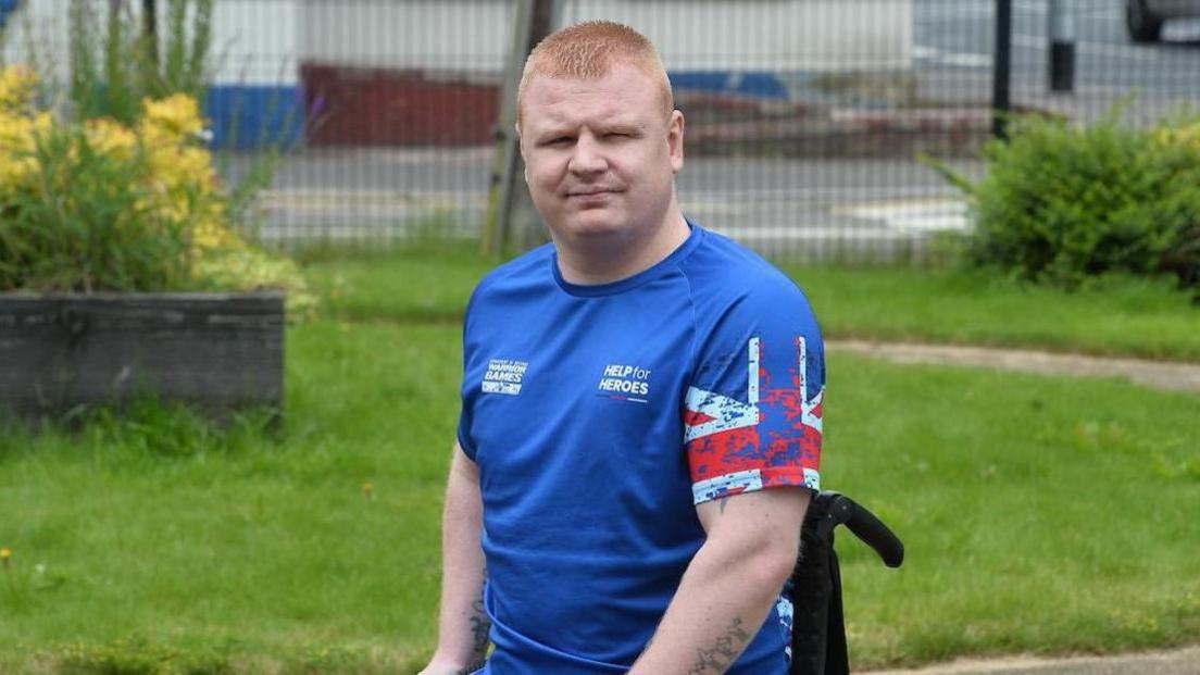 Andy Allen - a man is sitting in a wheelchair and is wearing a blue tshirt with 'help for heroes' on it. He is sitting in the middle of a garden and looking at the camera.