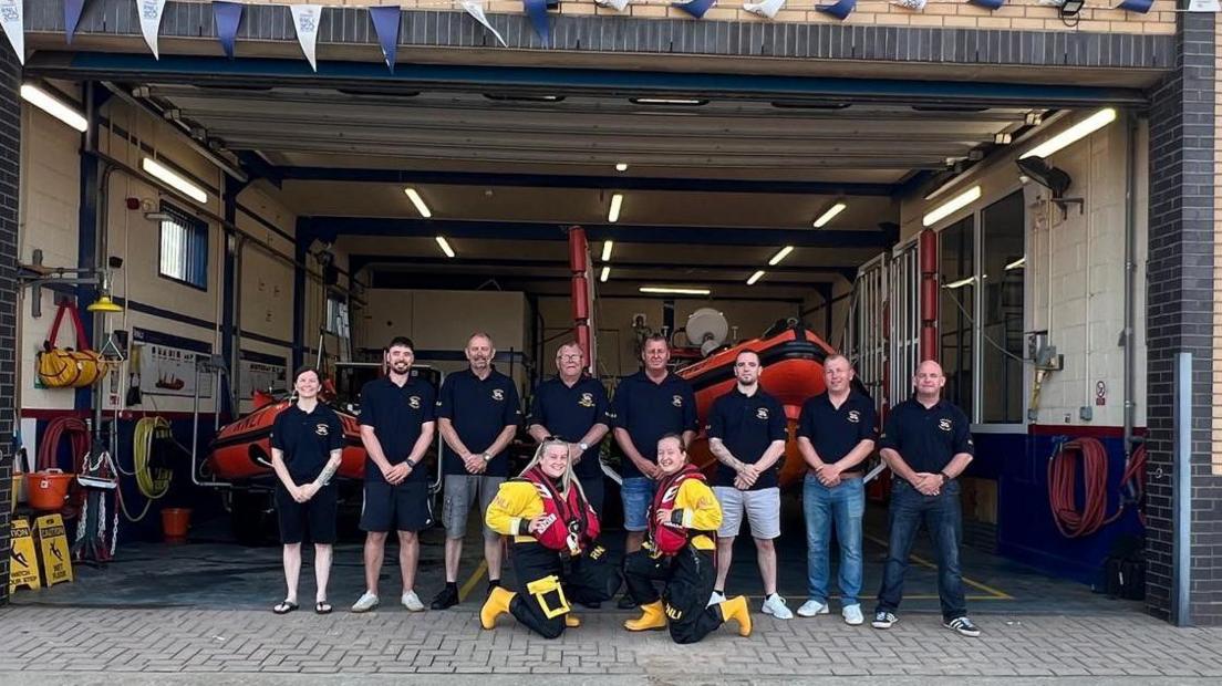 Members of the team standing in front of the lifeboat station wearing RNLI tops. Two of the team are crouched down in front dressed in yellow and black rescue gear